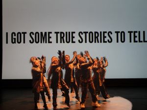Dancers clap their hands above their heads. In the background, a projection of the text "I got some true stories to tell"